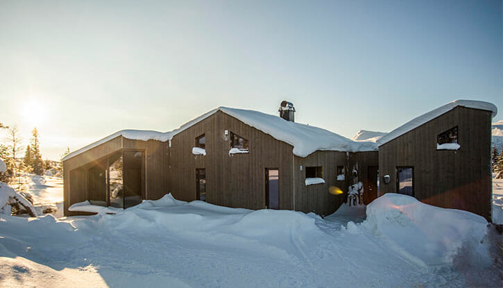 Foto. Moderne hytte med moderne vinkler og brun kledning. Snø på taket og på bakken i forgrunnen. Blå himmel og sol i bakgrunnen.