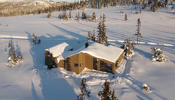 Dronefoto av hytte i vinterlandskap. Snø på taket og på bakken rundt hytta. Hytta er lysebrun.