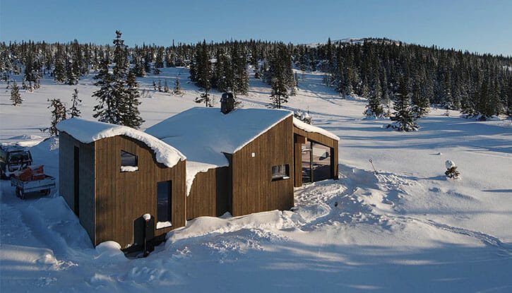 Foto av brun minimalistisk hytte i vinterlandskap. Snø omkring hytta og på taket. Trær i bakgrunnen. Blå himmel.