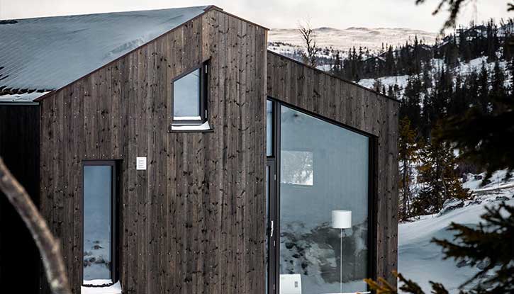 Foto av moderne mørkebrun hytte med skrå vinduer. Vinterlandskap i bakgrunnen. Foto.