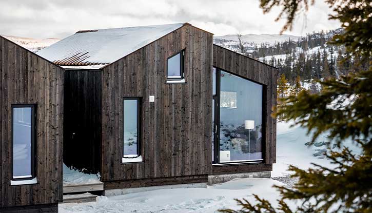 Foto av moderne mørkebrun hytte med skrå vinduer. Vinterlandskap i bakgrunnen. Foto.