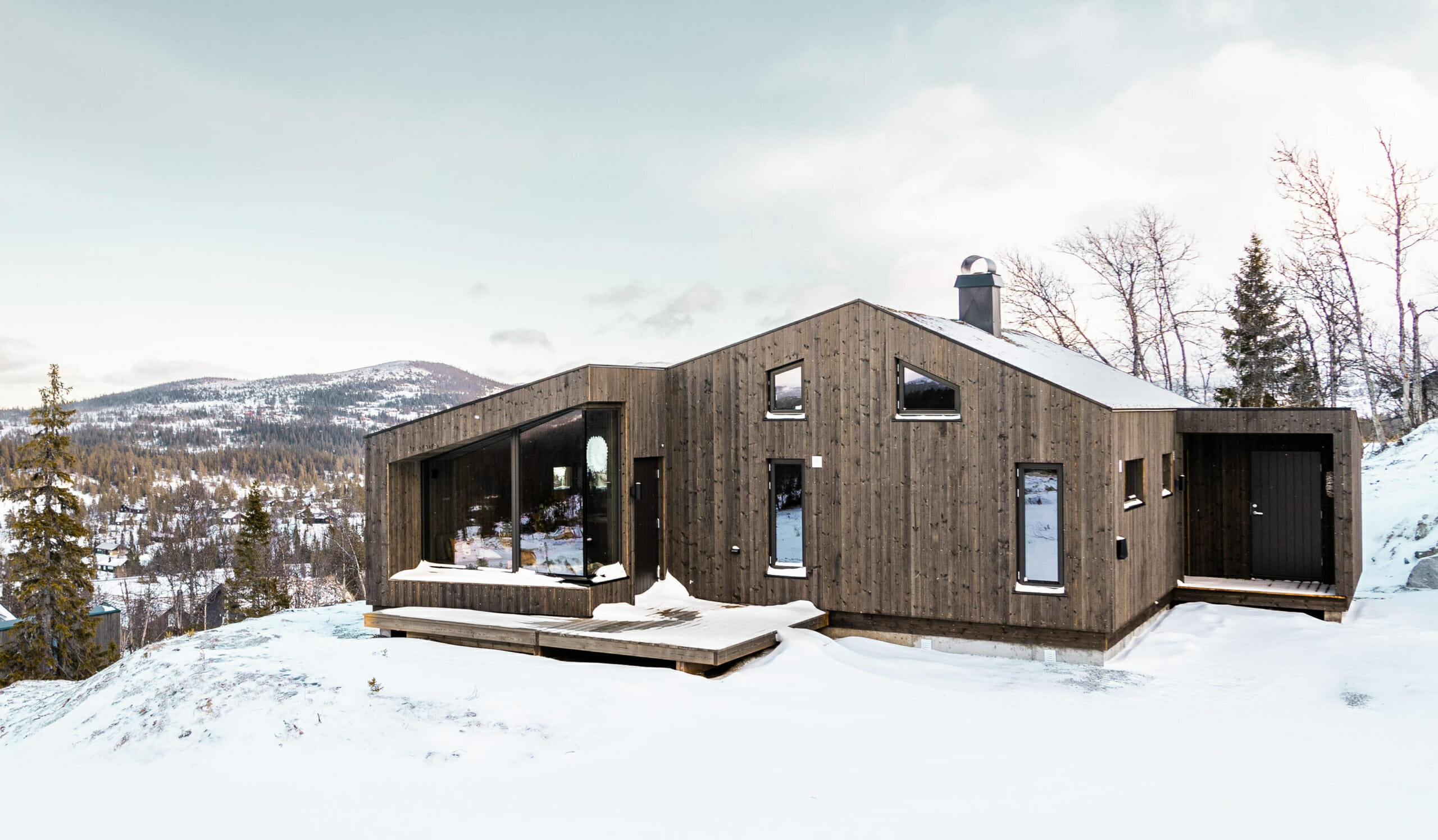 Foto av eksteriør hytte i gråbrun beiset kledning. Moderne hytte med vinduer med skrå vinkler. Snø på bakken og fjellandskap i bakgrunnen.