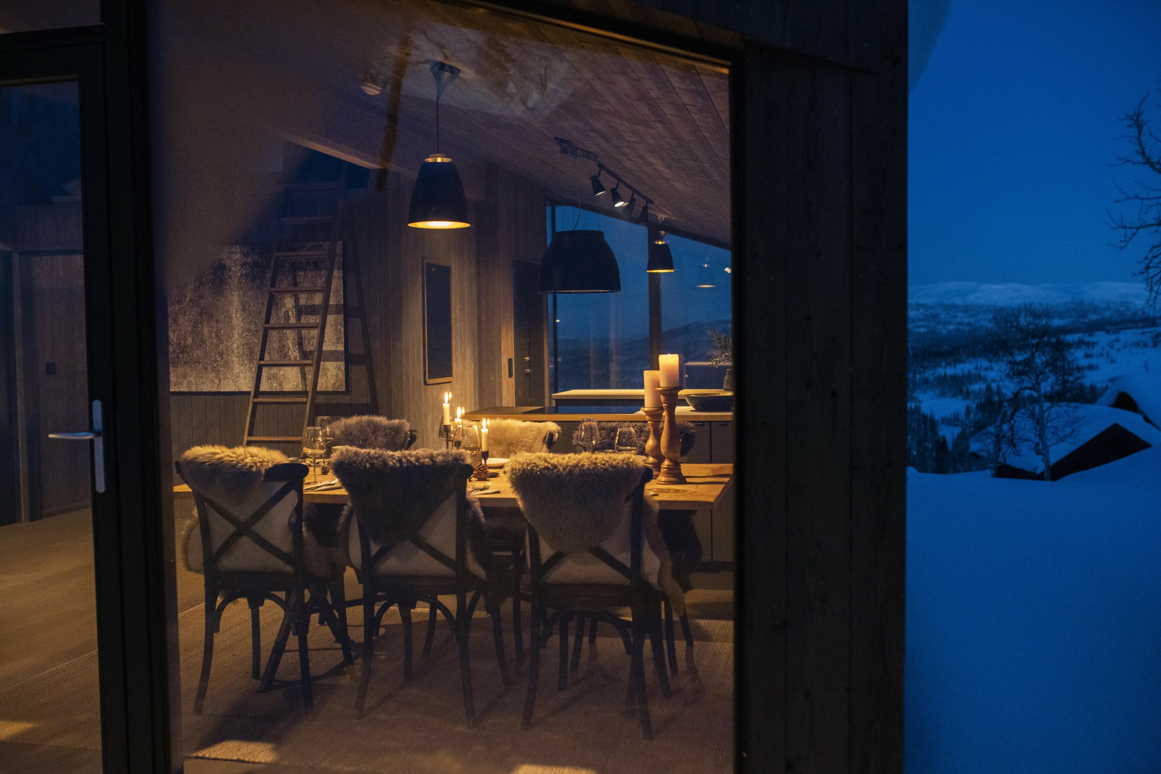 Spisestue i LY Hytta sett utenfra gjennom et stort vindu. Det er lyst og koselig inne, og blåtime med snø ute. Foto.