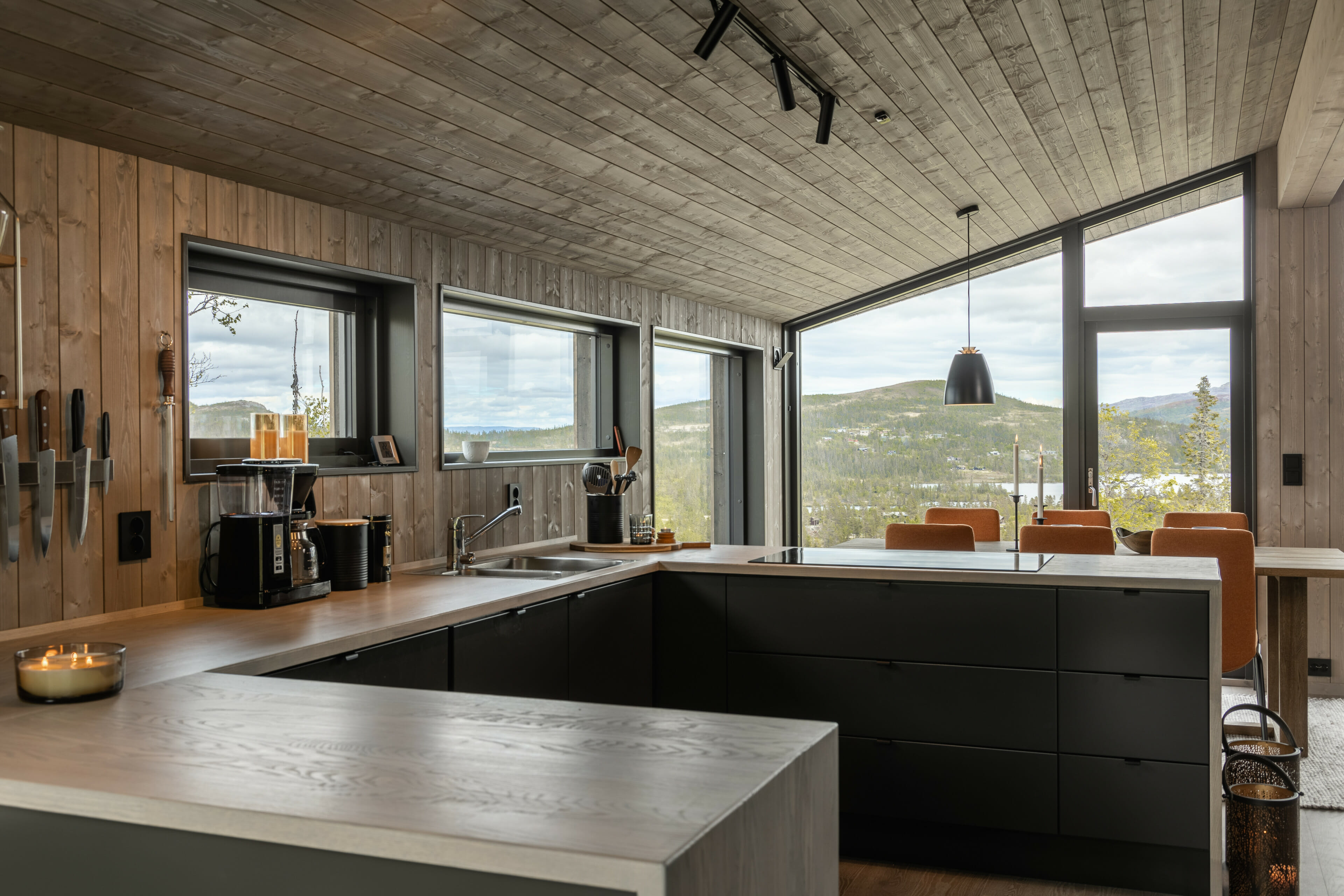 Foto. Interiør hytte med kjøkken og spisestuebord. Store vinduer med utsikt til fjell.