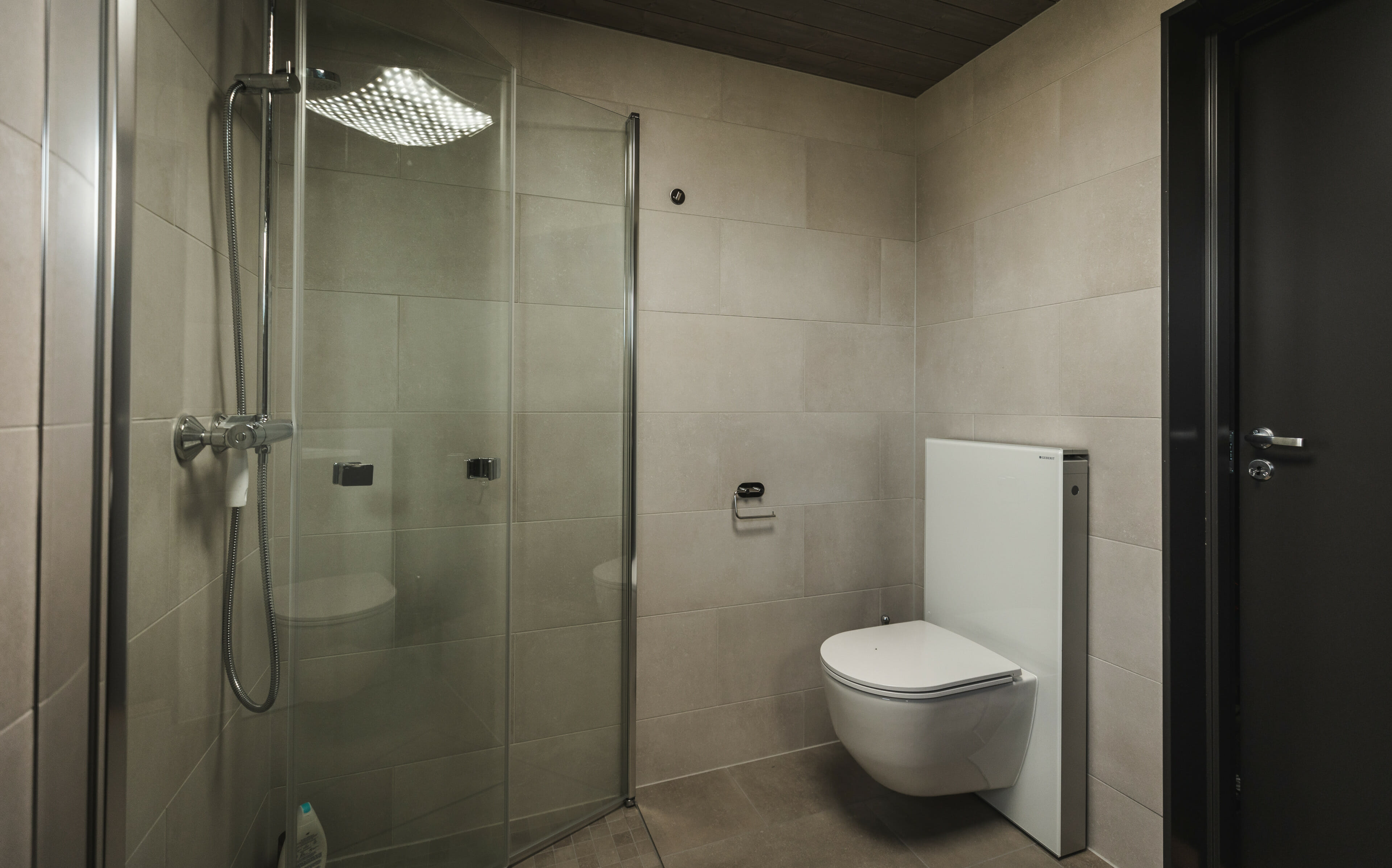 Foto. Interiør bad. Lyse fliser, vegghengt toalett og dushjørne med glassdører.