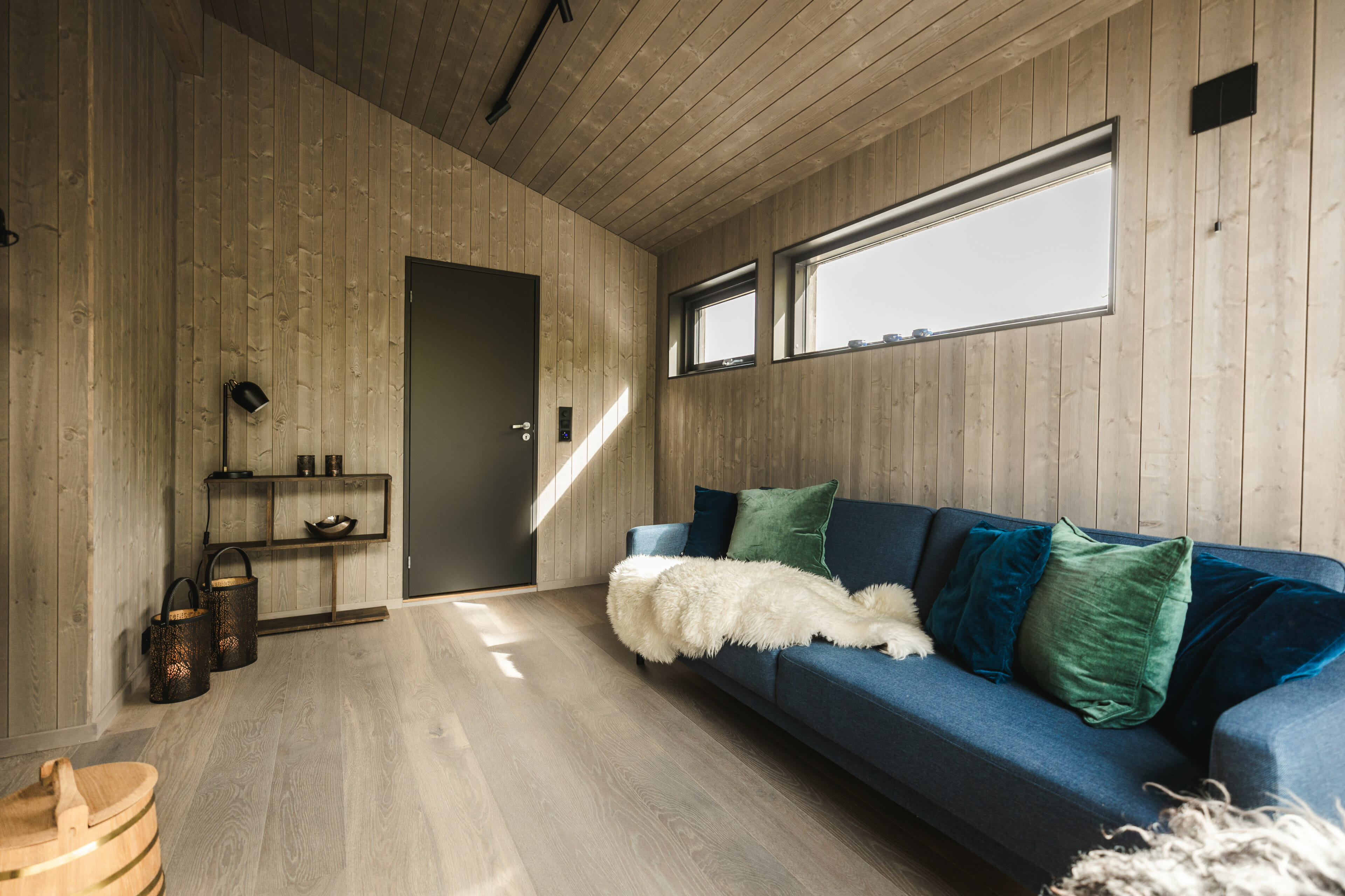 Foto innvendig av stue i moderne hytte. Trepanel på veggene. Blå sofa med grønne og blå puter.