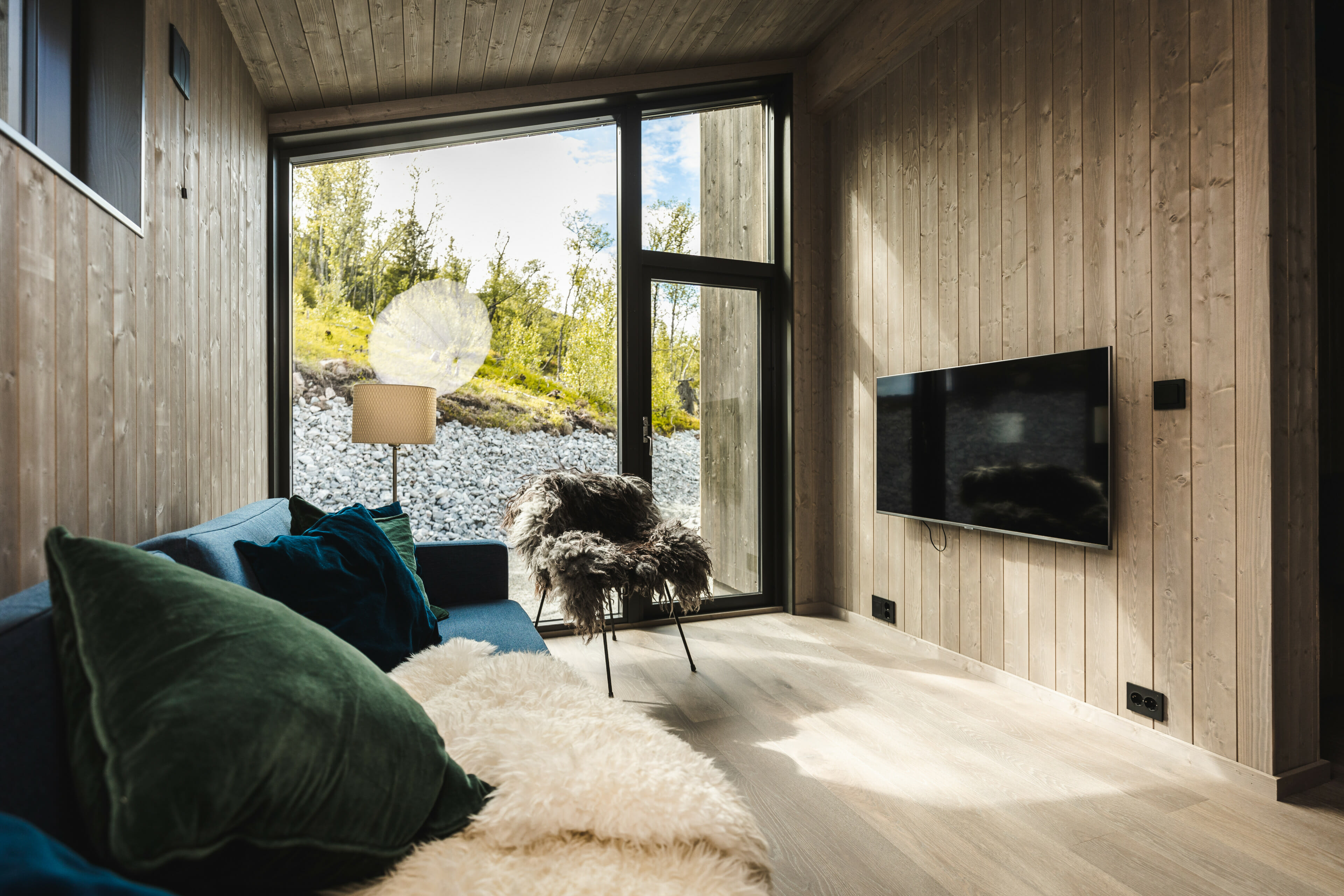 Interiørfoto fra stue i moderne hytte. Stort vindu og glassdør. Trepanel på veggene. Blå sofa med grønne og blå puter og vegghengt TV.