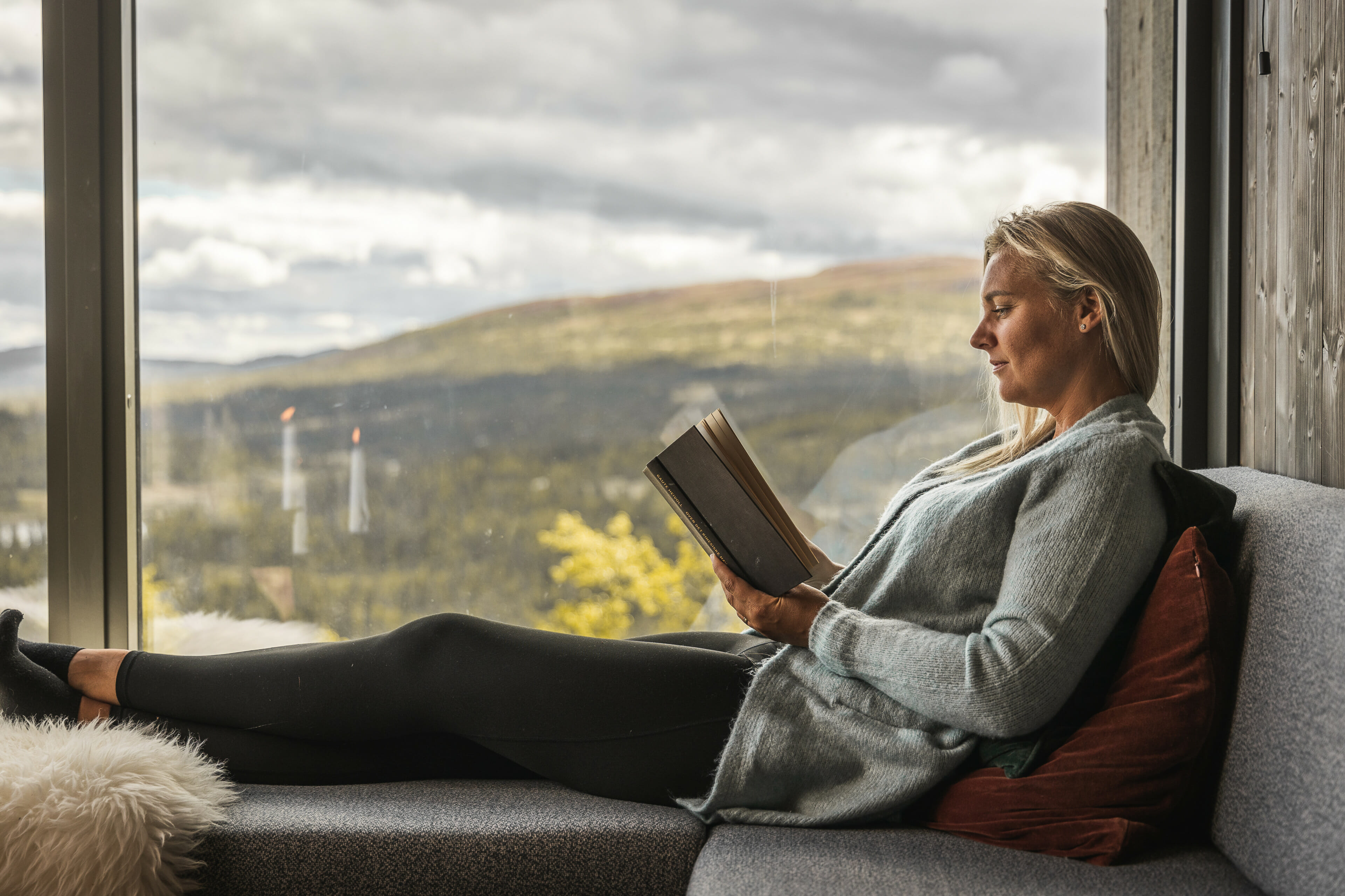 Kvinne i sofakrok inne i hytte leser bok. Stort vindu med utsikt til fjellandskap i bakgrunnen. Foto.