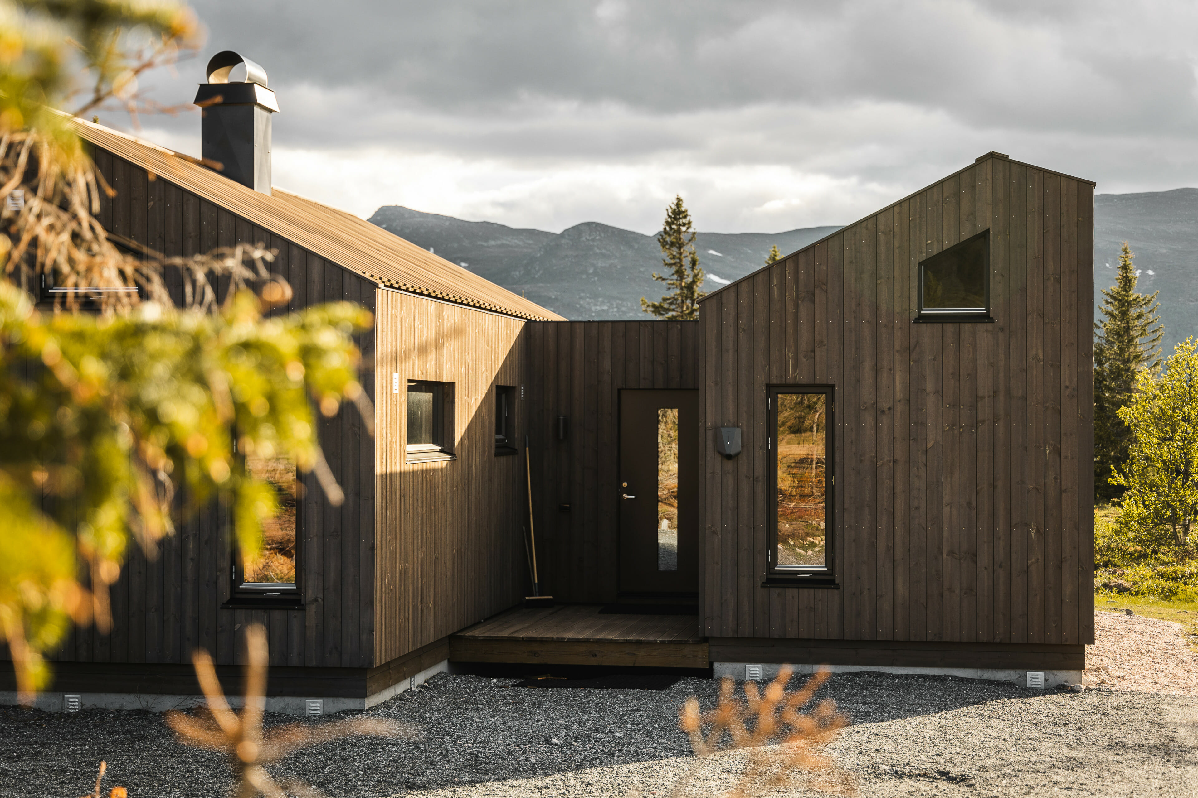 Foto. Eksteriør hytte med moderne vinkler. Minimalistisk design. Brun kledning. Høstfarger og fjellandskap i bakgrunnen.