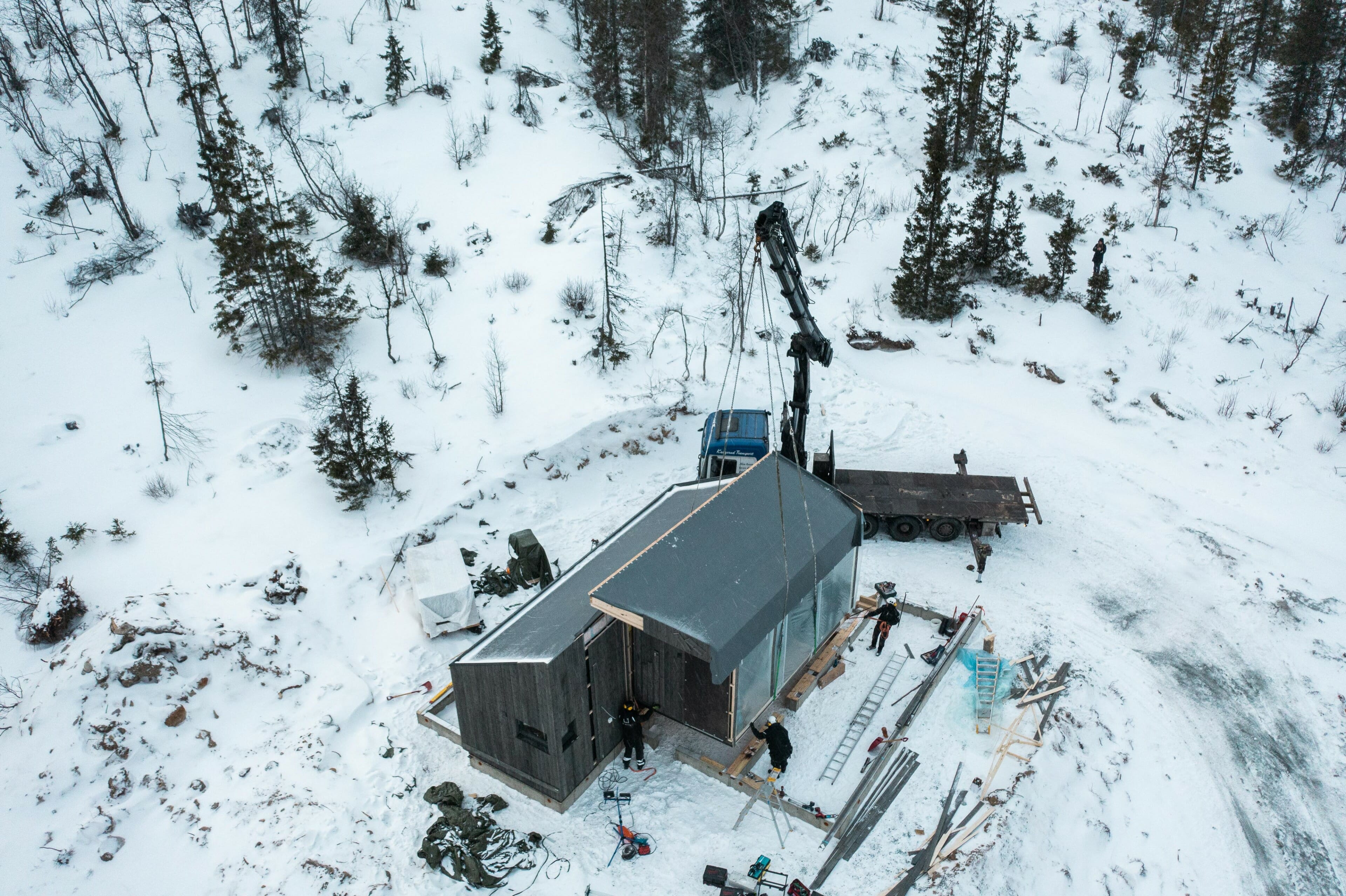 Dronefoto. Lastebil med heisekran plasserer element til hytte på tomt. Snø på bakken.