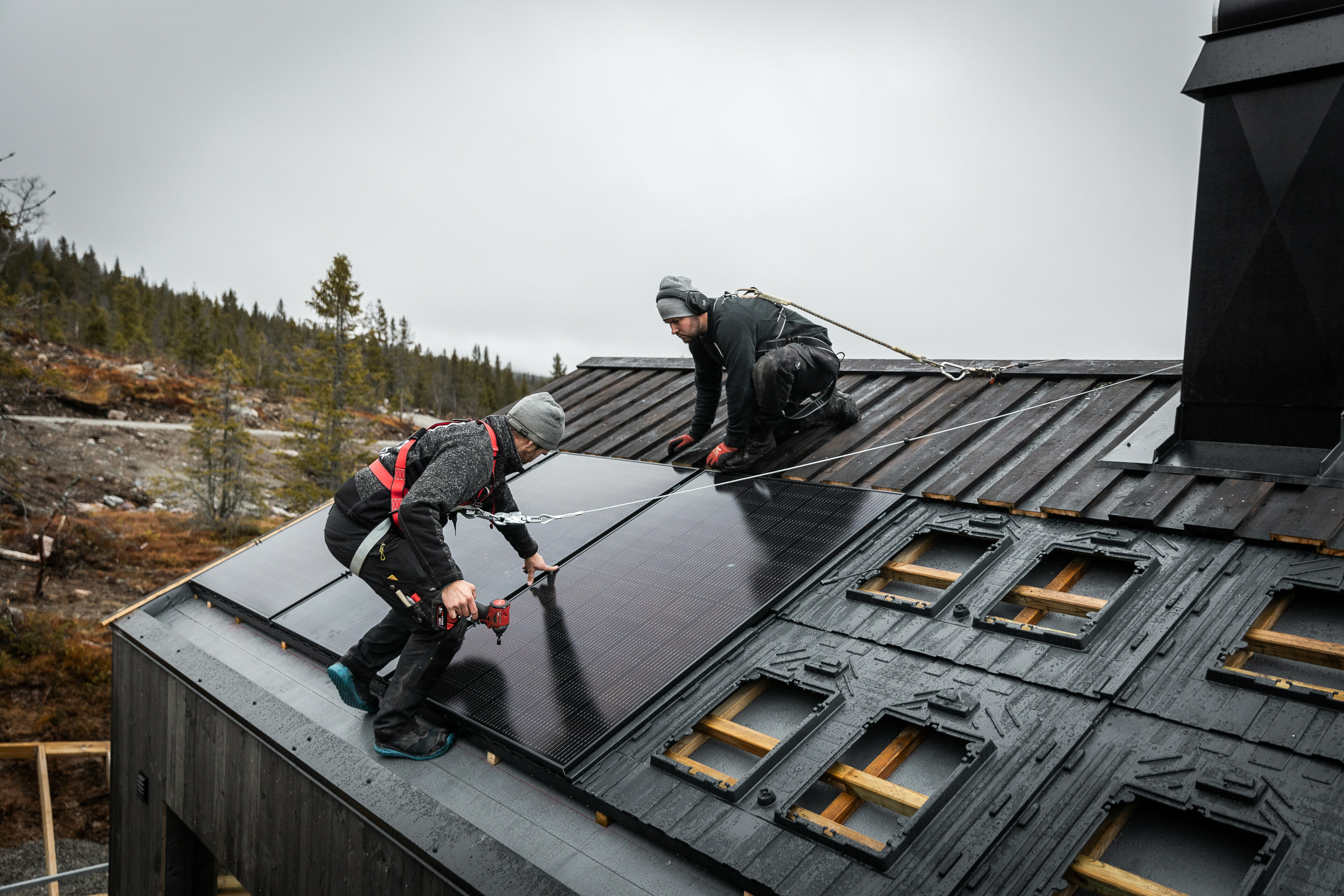 Hyttetak med to menn som arbeider med montering av solcellepanel. Mennene har sikkerhetsseler på seg. Foto.